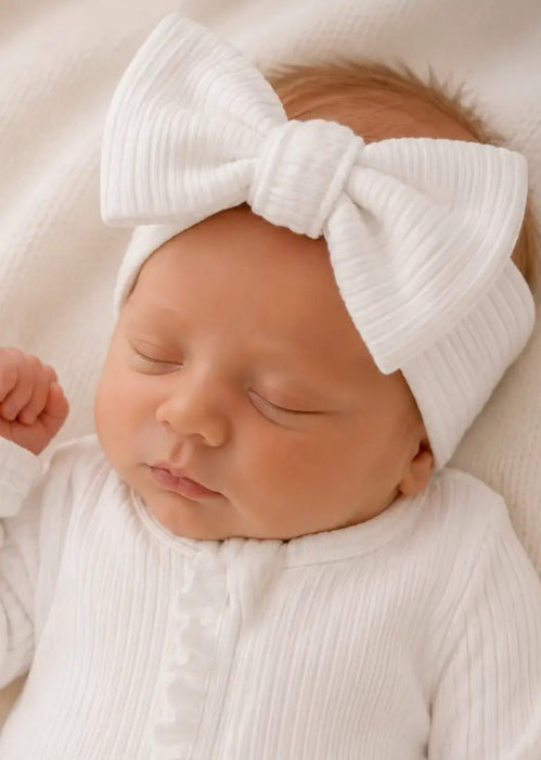A baby wearing a white ribbed headband with a bow, laying on a soft surface.
