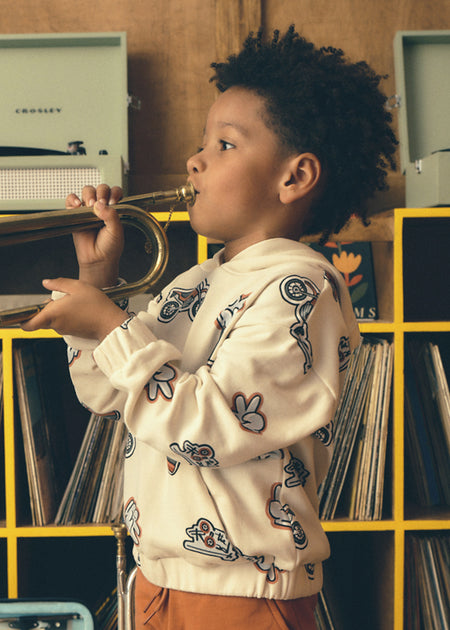 Child wearing a cream sweatshirt with a playful graphic print, playing a trumpet indoors.