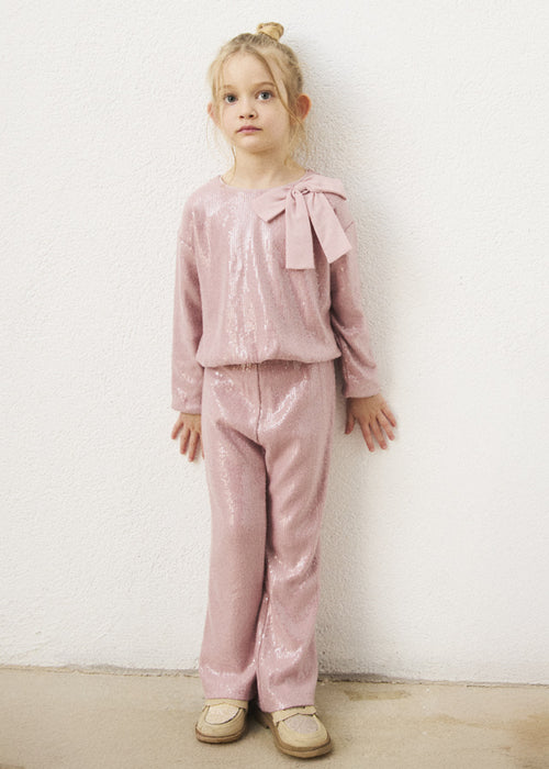 A toddler wearing a pink sparkle two-piece set with a large bow on the shoulder.
