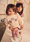 Two toddlers wearing a flower-embroidered sweatshirt and purple pants.