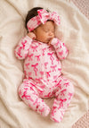 Baby wearing a pink footie with pink bows and a matching headband, lying on a soft blanket.