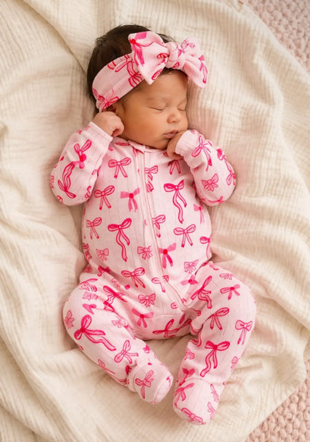 Baby wearing a pink footie with pink bows and a matching headband, lying on a soft blanket.