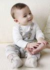 Baby wearing grey faux overalls with a dinosaur pattern, sitting on a cream couch.