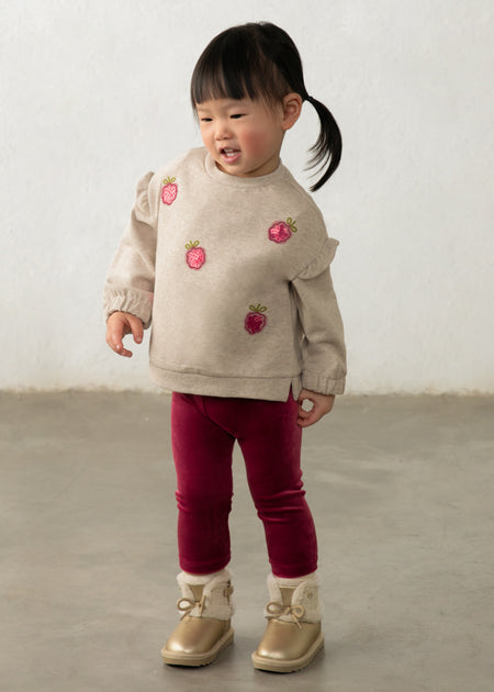 A toddler in a beige sweatshirt with raspberry appliqués and raspberry velour leggings.