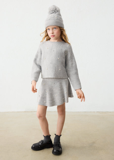A young girl wearing a grey knit sweater and skirt set with pearl embellishments, standing against a plain background.
