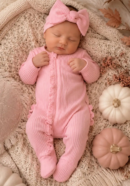 Baby in pink ribbed footie with frill detail and matching headband, lying on a knitted blanket.