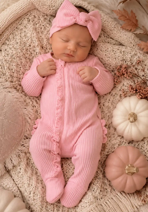 Baby in pink ribbed footie with frill detail and matching headband, lying on a knitted blanket.